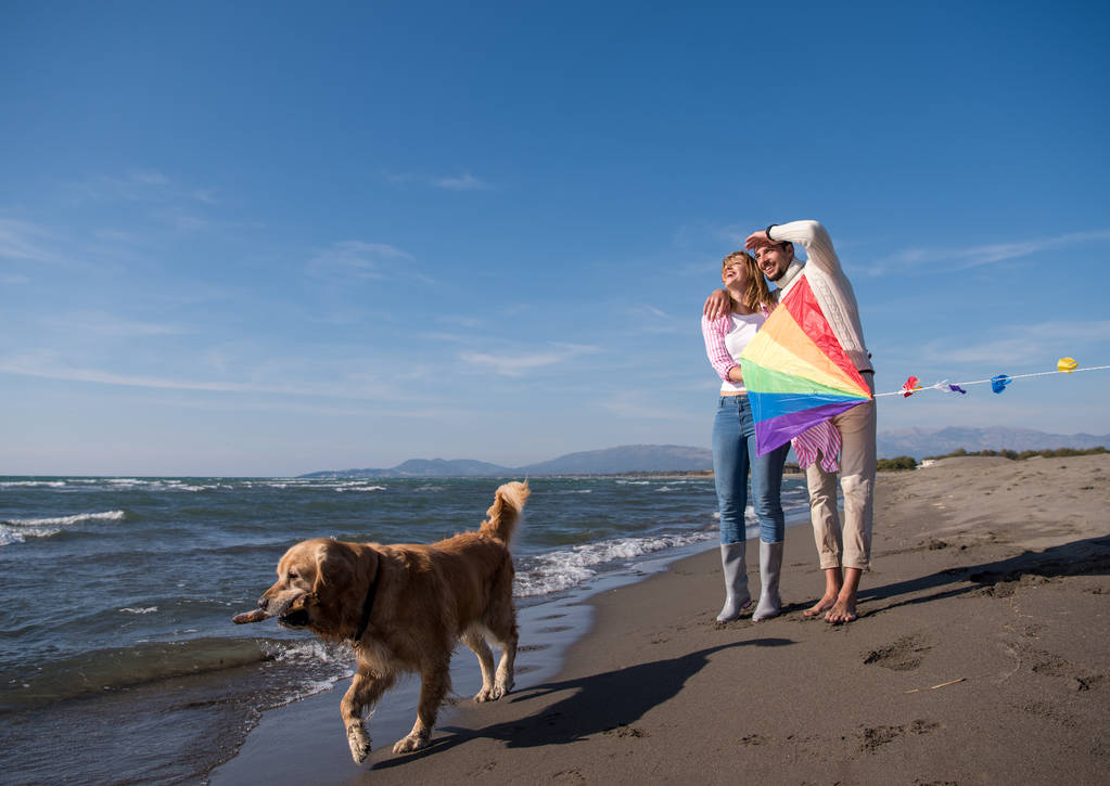 Casal jovem se divertindo brincando com um cachorro e pipa na praia no dia de outono
 - Foto, Imagem