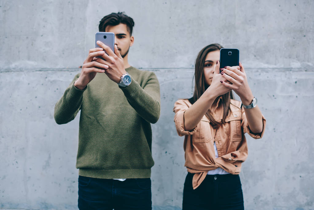Young teenagers holding mobile phones in front of their face concentrated on online chatting ignore real life communication, male and female friends addicted to modern smartphones standing on wall - Photo, Image