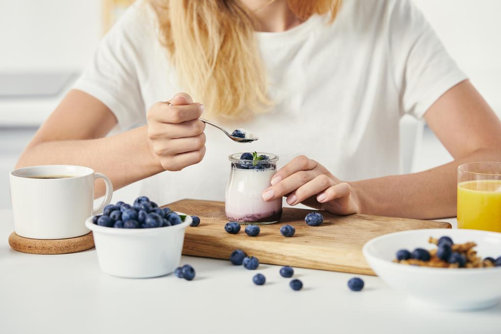abgeschnittene Aufnahme einer Frau an der Tischplatte mit Joghurt mit Blaubeeren, Tasse Kaffee und Glas Saft zum Frühstück - Foto, Bild
