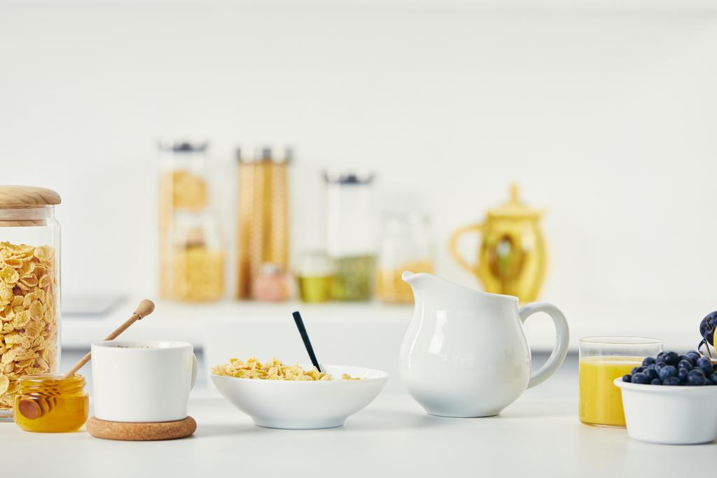 primer plano vista de copos de maíz en tazón, taza de café y jarra de leche para el desayuno en la superficie blanca
 - Foto, imagen