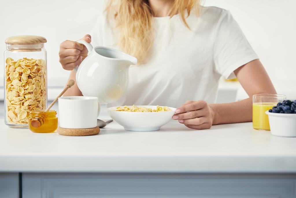 částečný pohled na ženu s džbán mléka sedí u stolu s miskou corn flakes a sklenici šťávy k snídani doma - Fotografie, Obrázek