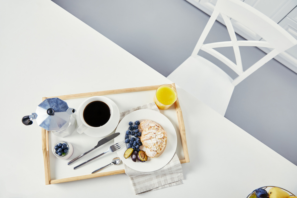 top view of croissant with fresh blueberries and plum pieces, glass of juice, cup of coffee and yogurt on wooden tray for breakfast on white tabletop - Photo, Image