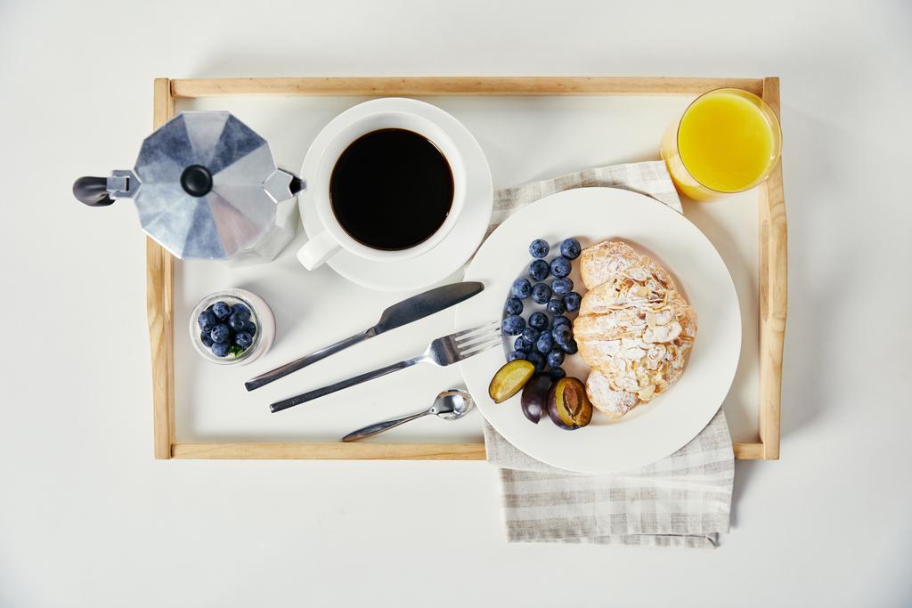 κάτοψη του κρουασάν με φρέσκα βακκίνια και δαμάσκηνο τεμάχια, ποτήρι χυμό, καφέ και γιαούρτι σε ξύλινη θήκη για πρωινό λευκά επιτραπέζια - Φωτογραφία, εικόνα