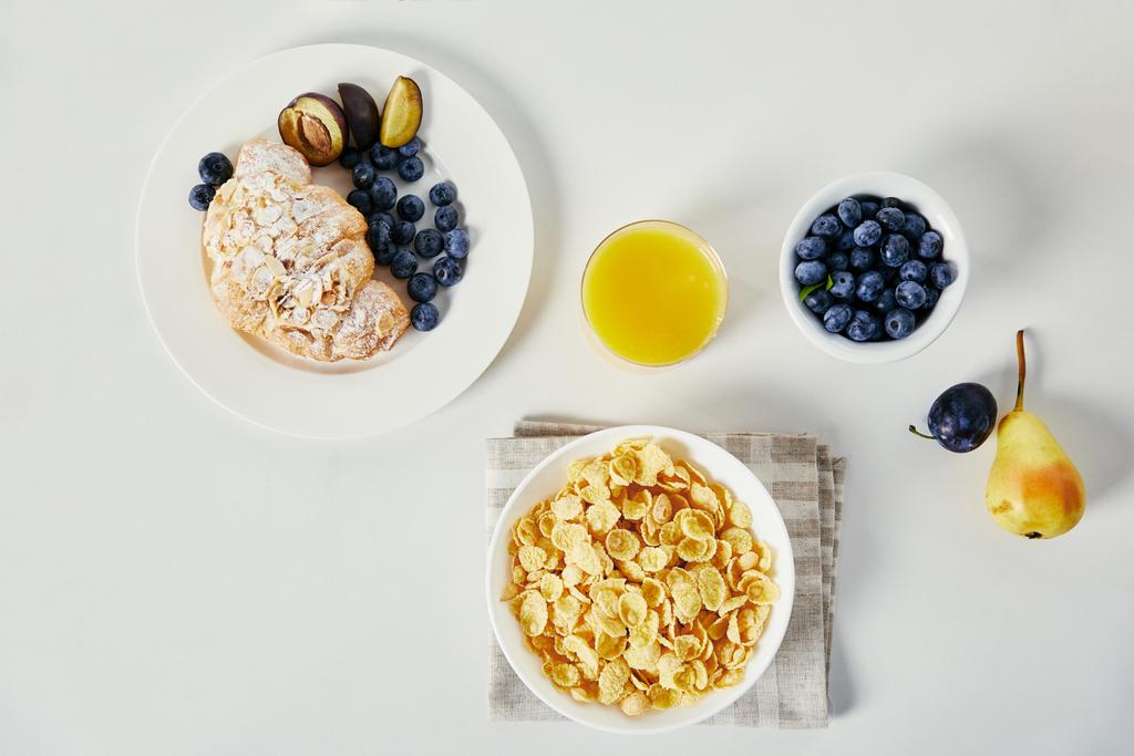 flat lay with corn flakes in bowl, glass of juice and croissant with blueberries and plum pieces for breakfast on white tabletop - Photo, Image
