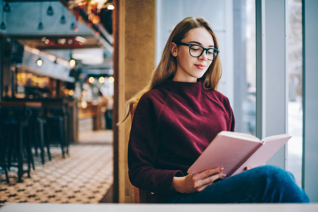 Sikeres diáklány viselnek a szemüveg a szakos egyetemi kávézó, kaukázusi intelligens csípő lány alkalmi élvezve látáskorrekcióra öltözött pozitív könyvével beltéri - Fotó, kép
