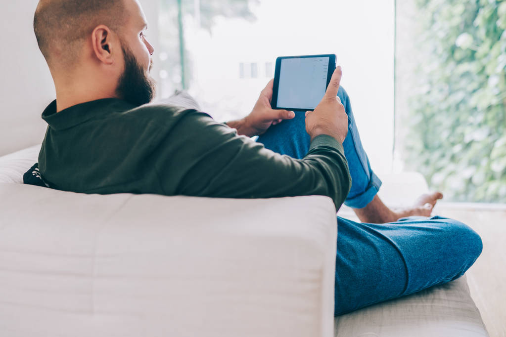 Вид сзади мужчины, лежащего на диване в квартире с помощью цифрового планшета для интернет-покупок, парня-хипстера, читающего интересную книгу на современном портативном компьютере, проводящего свободное время дома
 - Фото, изображение