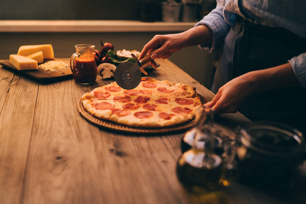 Женщина режет свежую запеченную домашнюю пиццу на деревенском кухонном фоне. Разрежьте на кусочки вкусную пиццу с грибами и ветчиной. Сыр и помидоры на деревянном столе. Здоровое питание, кулинарная концепция
. - Фото, изображение