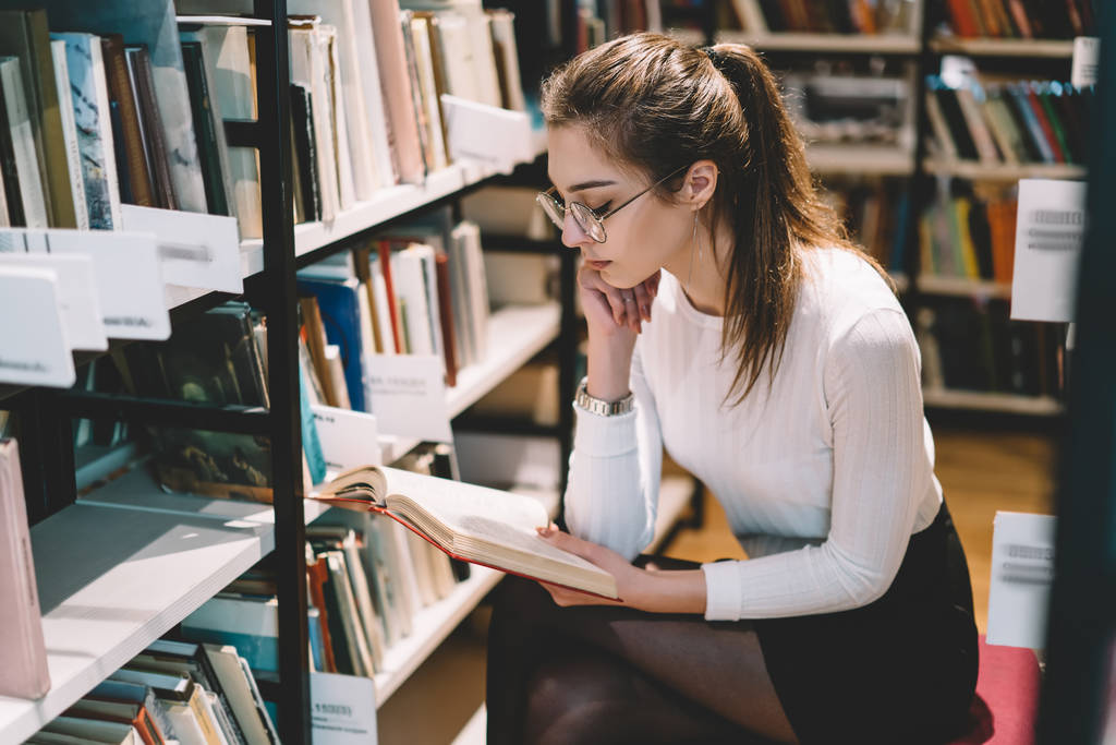 Pensive female student in eyewear for vision protection sitting near bookshelves reading interesting novel, smart concentrated young woman spending time in library fond of literature and educatio - Photo, Image