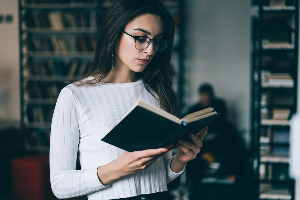 Mulher caucasiana em óculos da moda para correção de visão leitura de livros didáticos de aprendizagem para exames em biblioteca universitária, estudante concentrou-se em best-seller interessante gastar tempo em hobby
 - Foto, Imagem