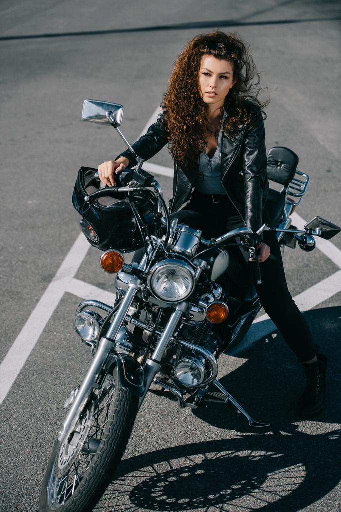 attraktives lockiges Mädchen sitzt auf klassischem Motorrad mit Helm auf dem Parkplatz - Foto, Bild
