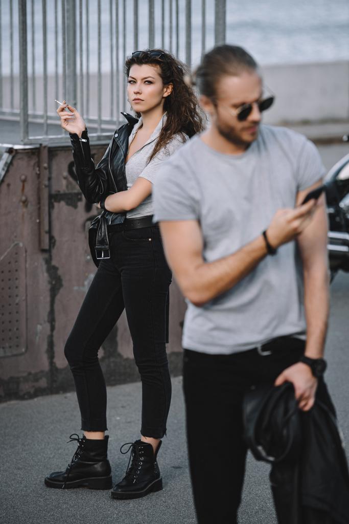 poikaystävä käyttää älypuhelinta, kun tyttöystävä seisoo klassisen moottoripyörän taustalla
 - Valokuva, kuva