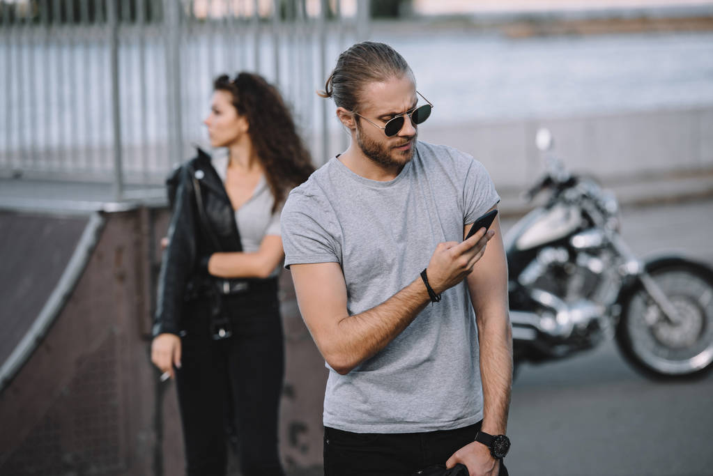 Mann benutzt Smartphone, während Mädchen mit klassischem Motorrad im Hintergrund steht - Foto, Bild