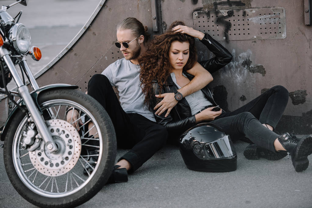 pareja de ciclistas abrazándose y sentados en asfalto con casco y motocicleta
 - Foto, imagen