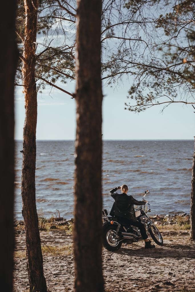rowerzysta w czarną skórzaną kurtkę, siedząc na motocykl klasyczny chopper, w pobliżu rzeki w lesie - Zdjęcie, obraz