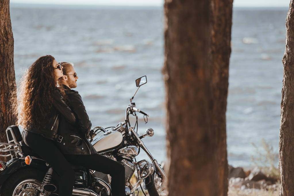 пара байкеров в черных кожаных куртках, сидящих на мотоцикле и смотрящих на море
 - Фото, изображение