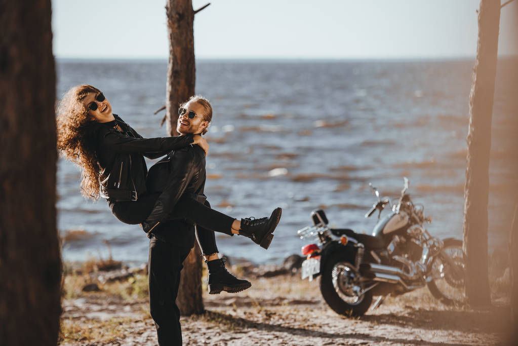novio divirtiéndose y sosteniendo a su novia en brazos en la orilla del mar con la motocicleta cerca
 - Foto, imagen