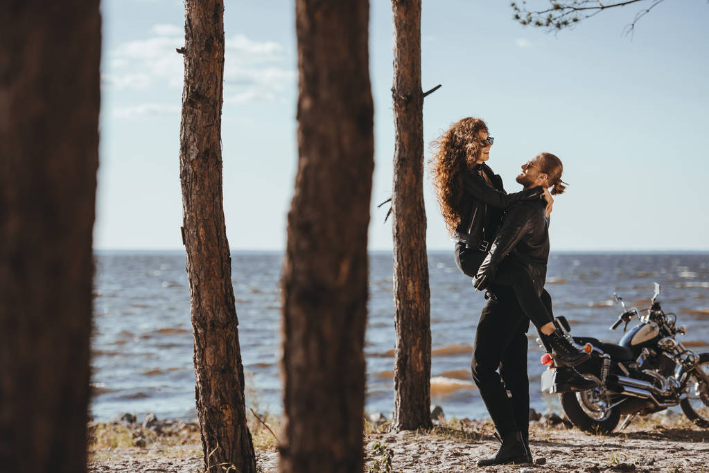 Freund hält seine glückliche Freundin am Strand mit Motorrad in der Nähe - Foto, Bild