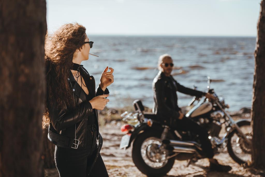seçici odak sigara ve sahil üzerinde motosiklet üzerinde erkek arayan kadın - Fotoğraf, Görsel