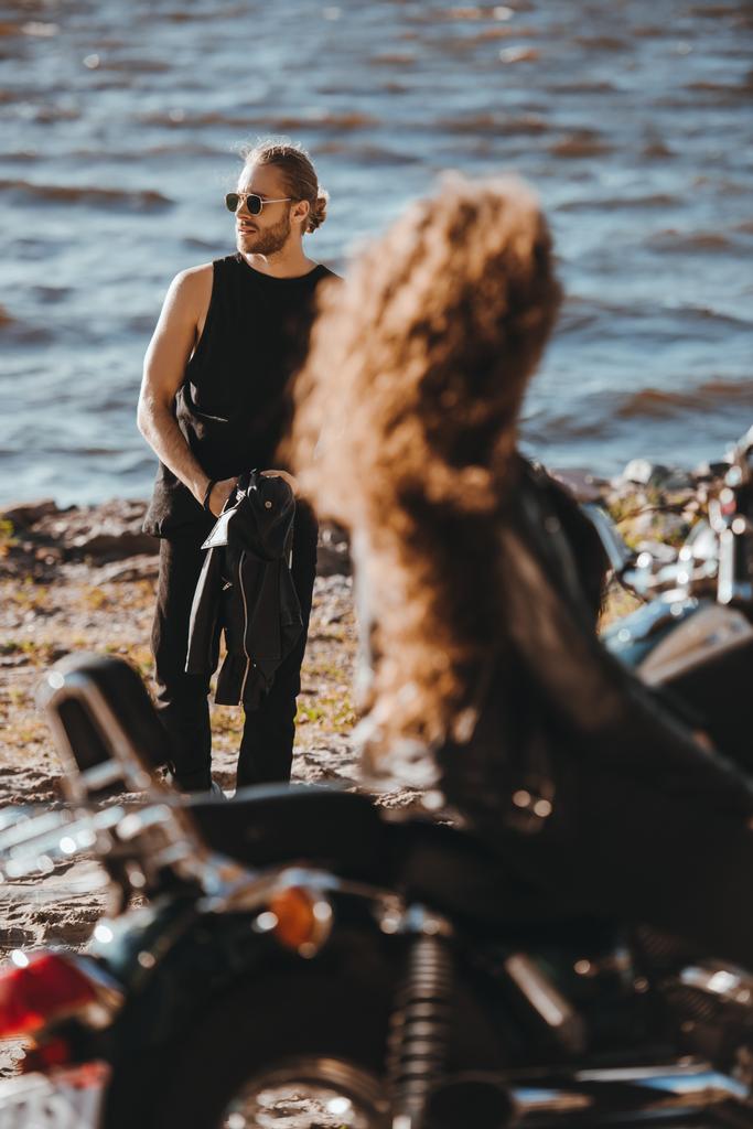 enfoque selectivo de la chica sentada en la motocicleta y mirando a su novio caminando en la orilla del mar
 - Foto, Imagen
