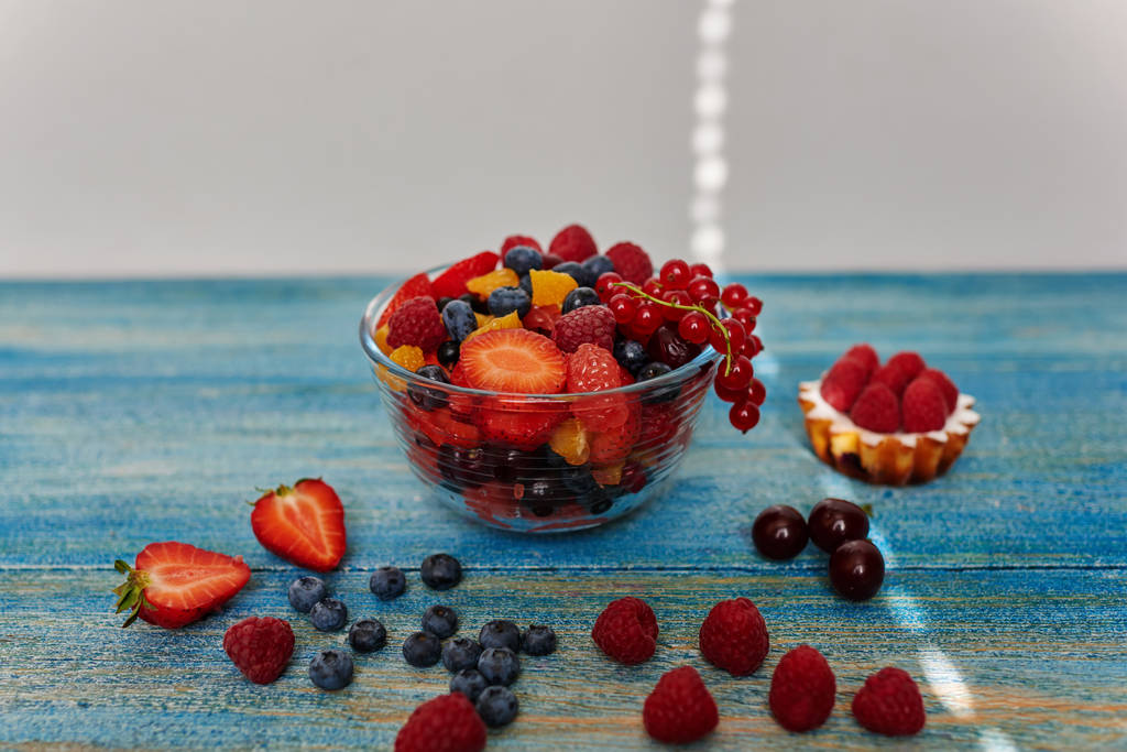 На столі вінтажне блакитне глибоке скляне блюдо зі свіжими нарізаними фруктами, поруч з невеликим тістом з тістечка зі свіжою малиною та домашнім джемом
 - Фото, зображення