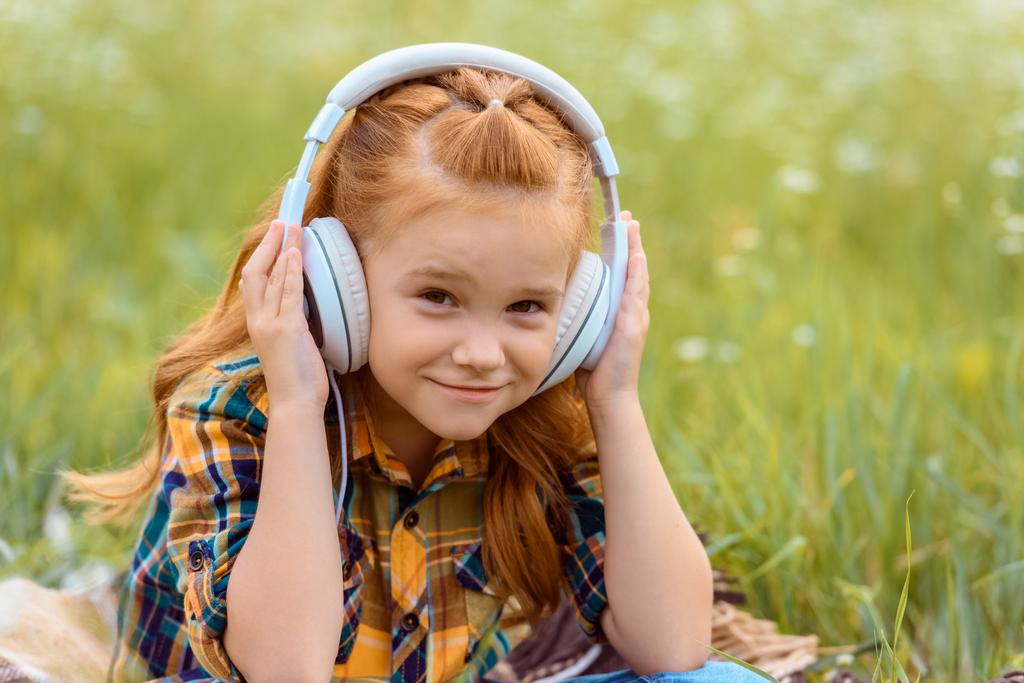 Porträt eines lächelnden Kindes, das über Kopfhörer Musik hört, mit grünem Gras auf dem Hintergrund - Foto, Bild