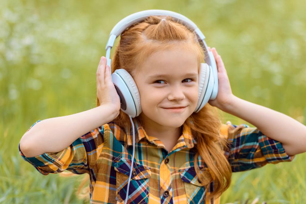 Portret van een glimlachend kind luisteren muziek in hoofdtelefoon met groen gras op achtergrond - Foto, afbeelding