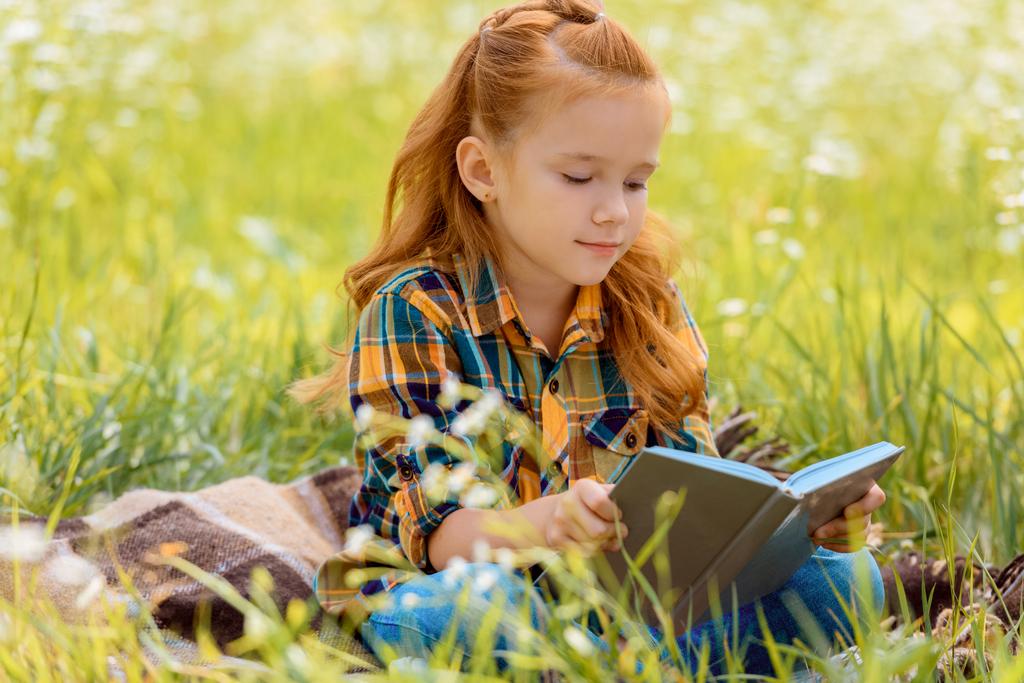 portrait of adorable red hair child reading book while sitting in summer field - Photo, Image