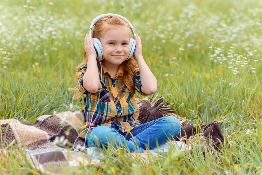 cute kid listening music in headphone while sitting on blanket in field with wild flowers - Photo, Image