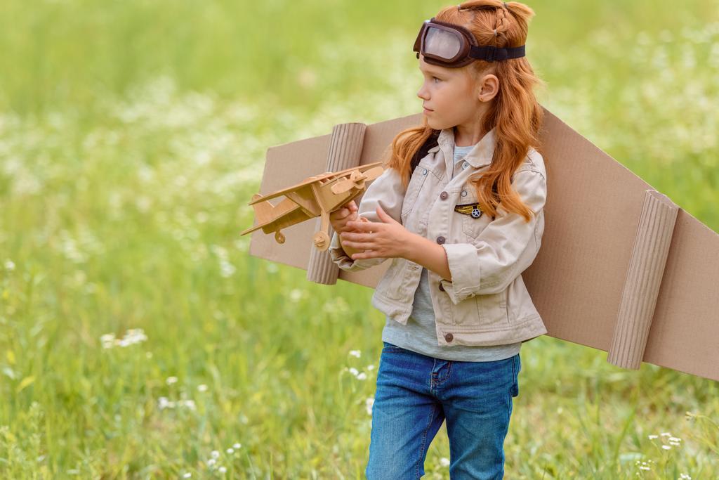 Porträt eines kleinen Kindes im Pilotenkostüm mit hölzernem Spielzeugflugzeug, das auf einer Wiese steht - Foto, Bild