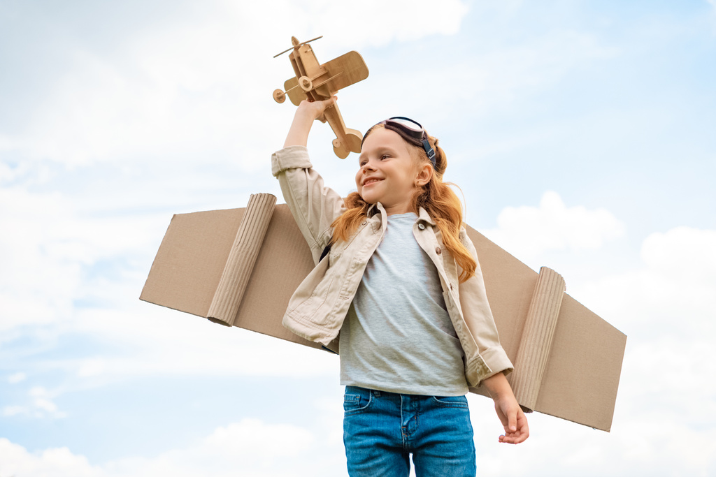 ローアングル パイロットの衣装を青い曇り空を背景に木製のおもちゃの飛行機を保持における子供観 - 写真・画像