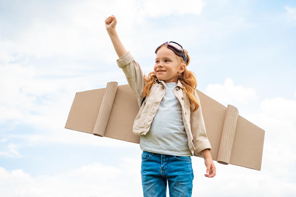 portrait of child with paper plane wings on back and protective eyeglasses on head standing with outstretched arm against blue cloudy sky - Photo, Image