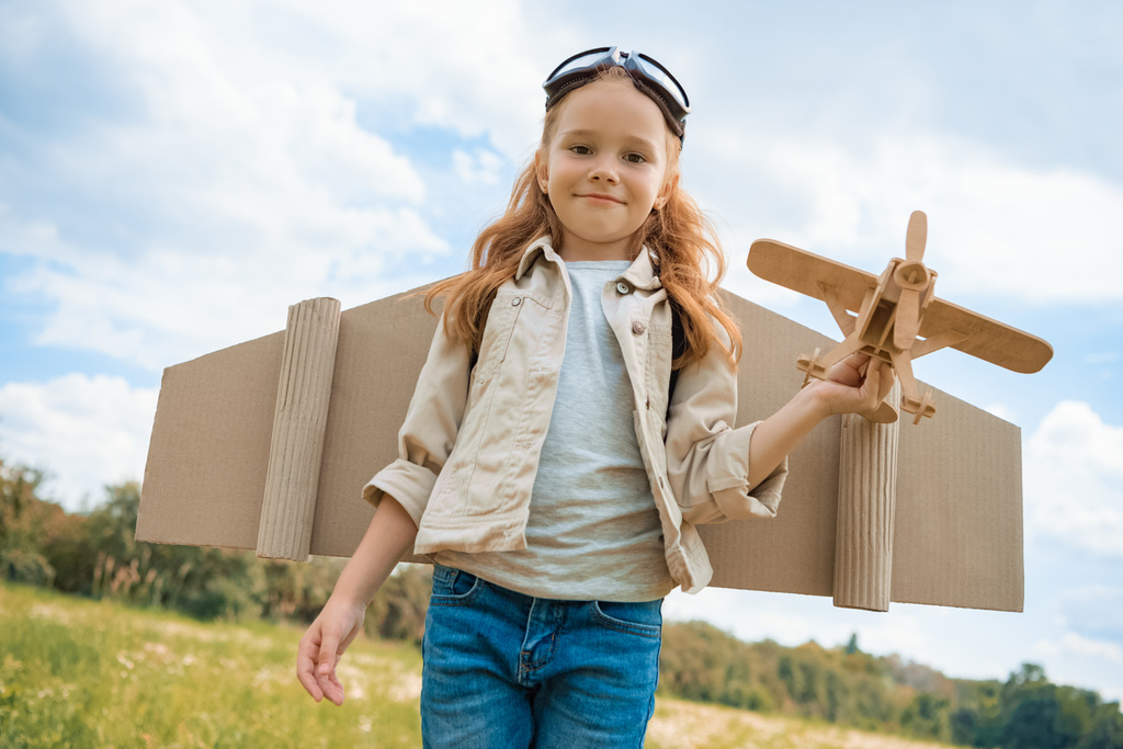 Porträt eines lächelnden Rothaarkindes im Pilotenkostüm, das ein Holzflugzeug im Sommerfeld hält - Foto, Bild