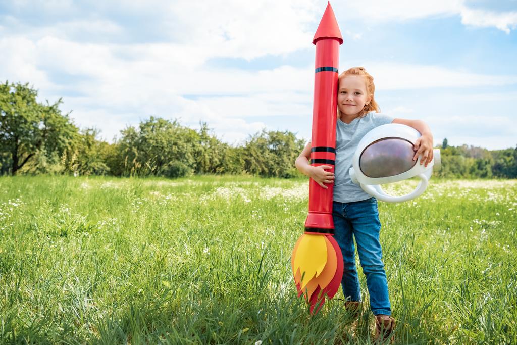 улыбающийся рыжий ребенок с ракетно-космическим шлемом, стоящий на летнем поле
 - Фото, изображение