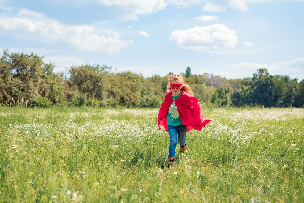 klein kind in rode superheld cape en masker in de weide op zomerdag uitgevoerd - Foto, afbeelding