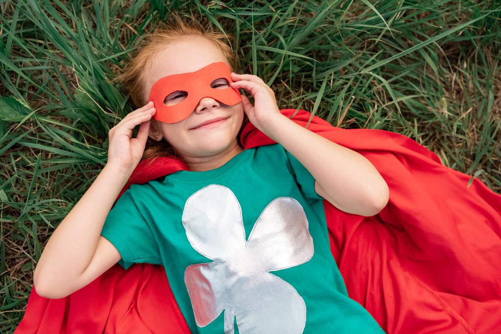 Blick von oben auf Kind in rotem Superheldenumhang und Maske, das auf grünem Gras liegt - Foto, Bild