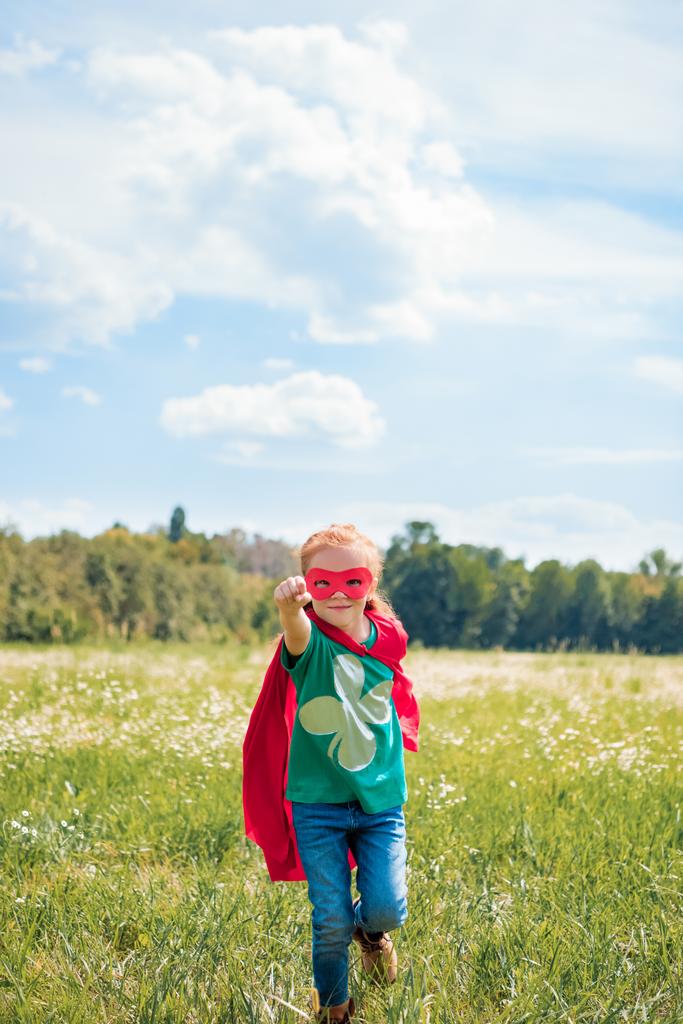 Kleines Kind in rotem Superheldenumhang und Maske mit ausgestrecktem Arm läuft an einem Sommertag auf der Wiese - Foto, Bild