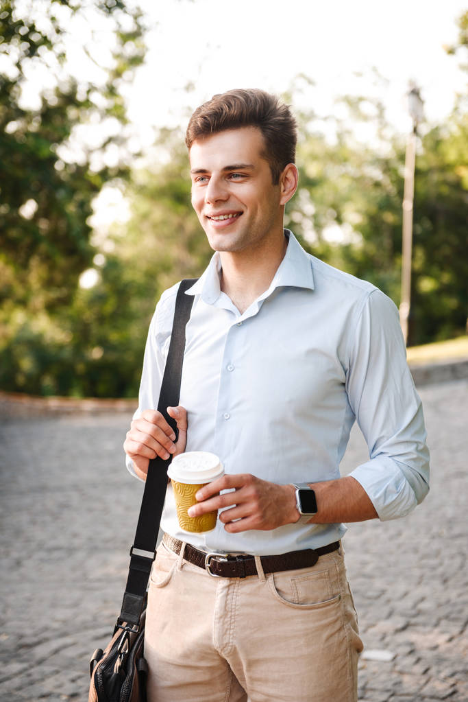 Щасливий молодий чоловік у сорочці, що несе сумку, що йде в міському парку з чашкою кави
 - Фото, зображення