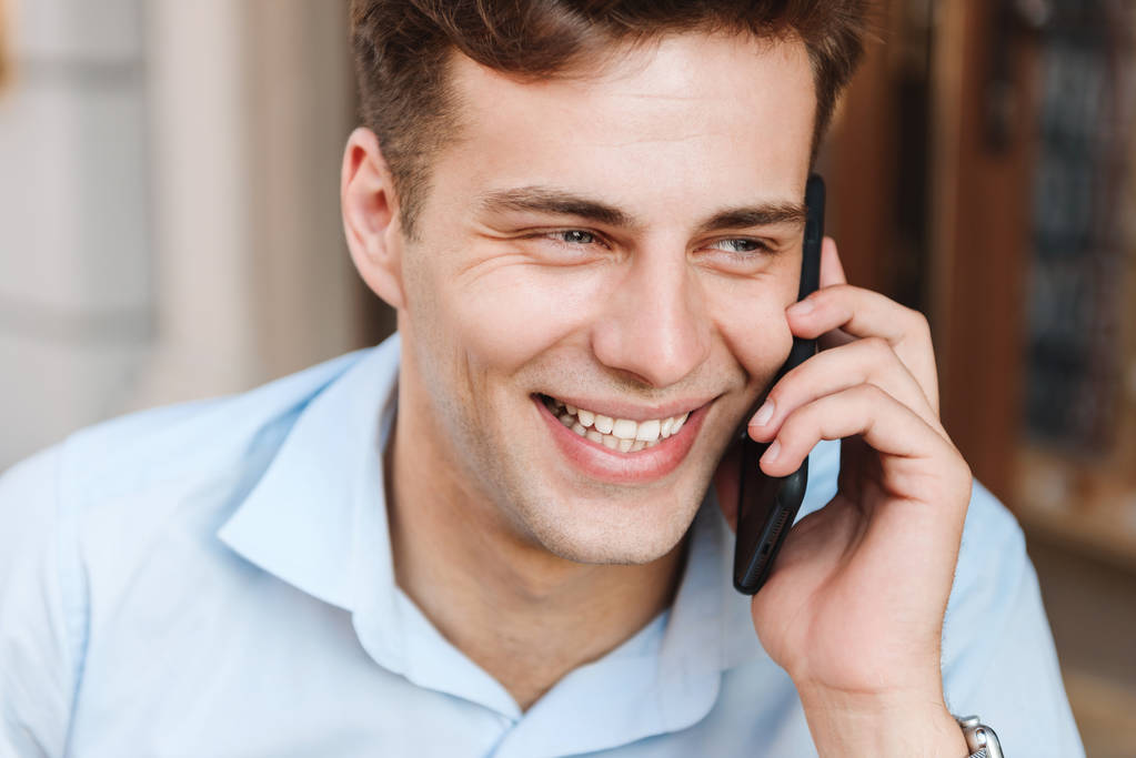 Portret van een lachende jongeman in shirt praten op mobiele telefoon buitenshuis close-up - Foto, afbeelding