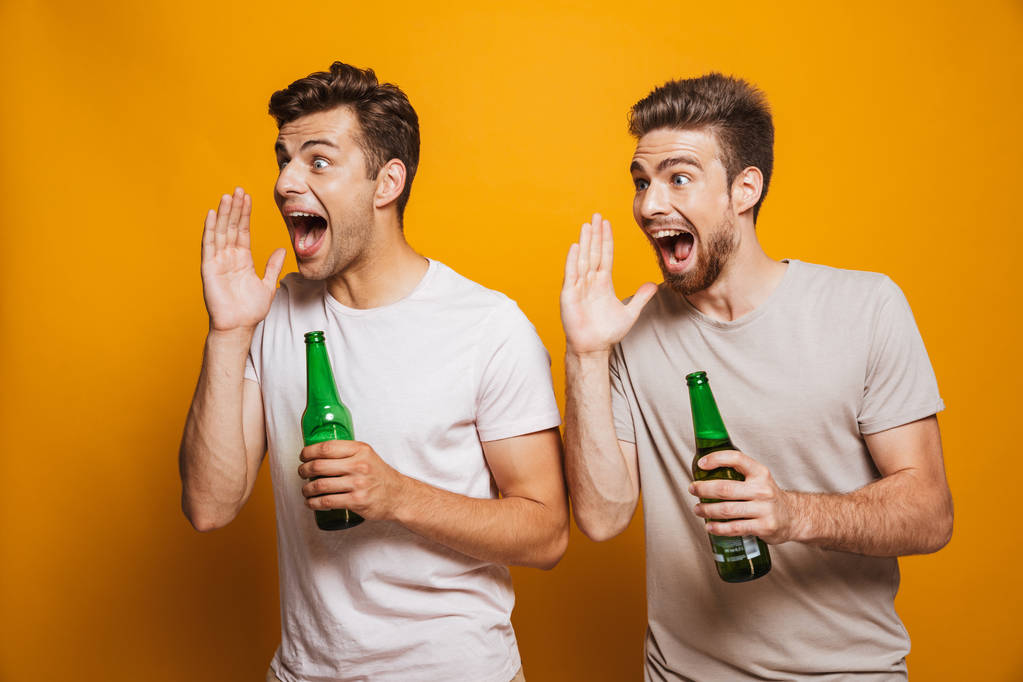 Портрет двух счастливых молодых людей, лучших друзей, держащих пивные бутылки и кричащих изолированные на желтом фоне
 - Фото, изображение