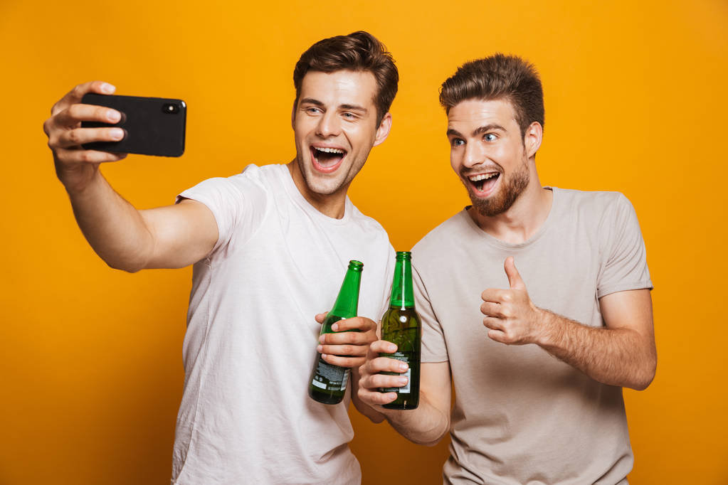 Portret van een twee vrolijke jonge mannen beste vrienden nemen van een selfie tijdens bedrijf bierflesjes en geïsoleerde schreeuwen over gele achtergrond - Foto, afbeelding