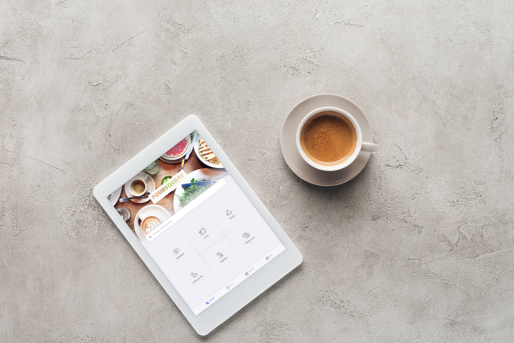 Draufsicht auf Tasse Kaffee und Tablet mit Foursquare-App auf Bildschirm auf Betonoberfläche - Foto, Bild