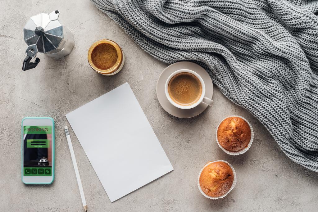 top view of cup of coffee with sweet muffins, blank paper and smartphone with booking app on screen on concrete surface with knitted wool drapery - Photo, Image