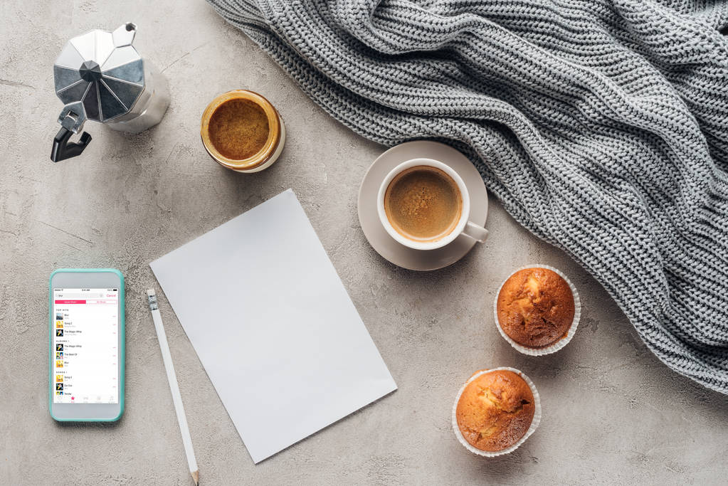 top view of cup of coffee with muffins, blank paper and smartphone with music playlist on screen on concrete surface with knitted wool drapery - Photo, Image