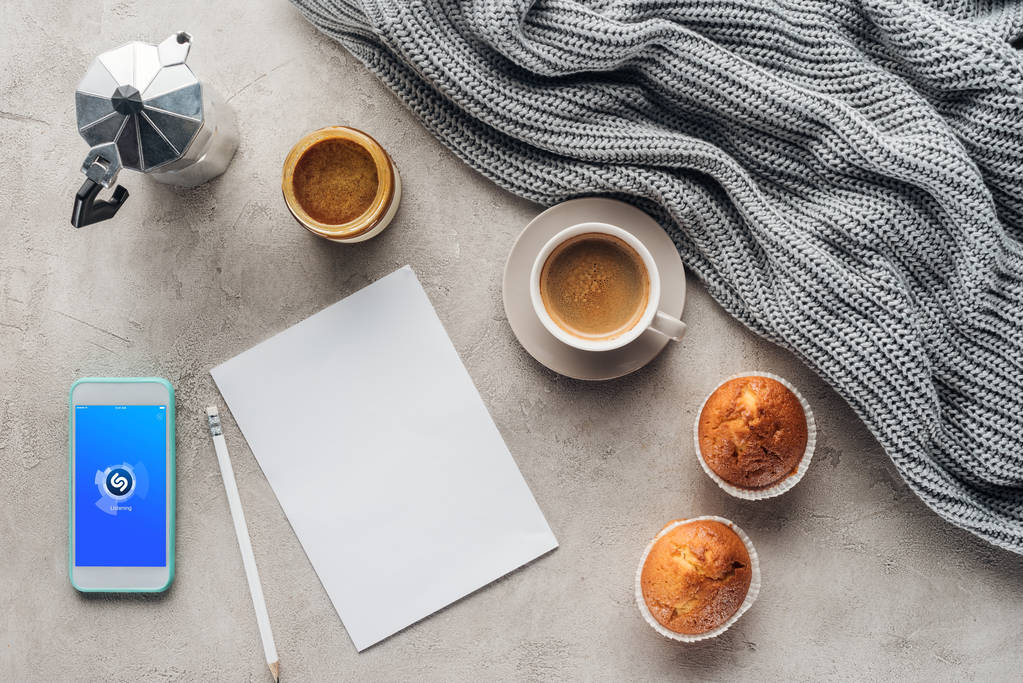 bovenaanzicht van een kopje koffie met muffins, blanco papier en smartphone met shazam app op scherm op betonnen oppervlak met gebreide wol gordijnen - Foto, afbeelding