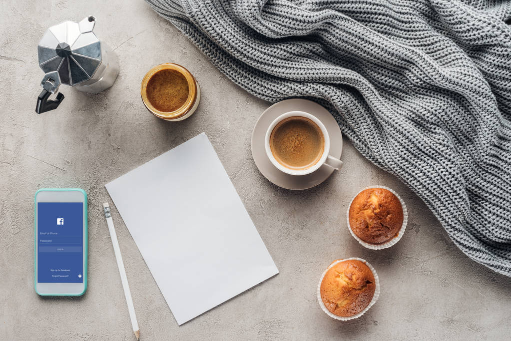 ylhäältä näkymä kuppi kahvia muffinsseja, tyhjä paperi ja älypuhelin facebook sovellus ruudulla betonipinnalla neulotulla villakankaalla
 - Valokuva, kuva