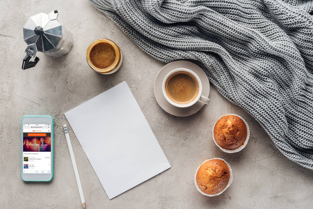 Draufsicht auf Kaffeebecher mit Muffins, leerem Papier und Smartphone mit Soundcloud-App auf dem Bildschirm auf Betonoberfläche mit Strickwolltuch - Foto, Bild