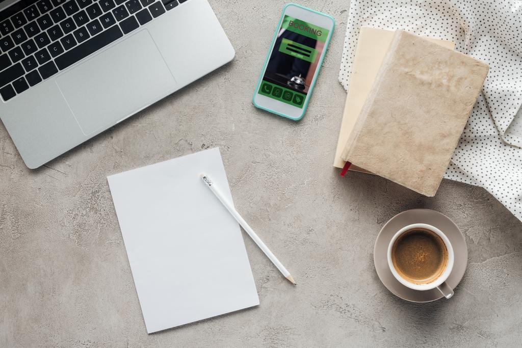 bovenaanzicht van koffie met laptop en smartphone met boeken app op scherm op betonnen oppervlak met blanco papier - Foto, afbeelding