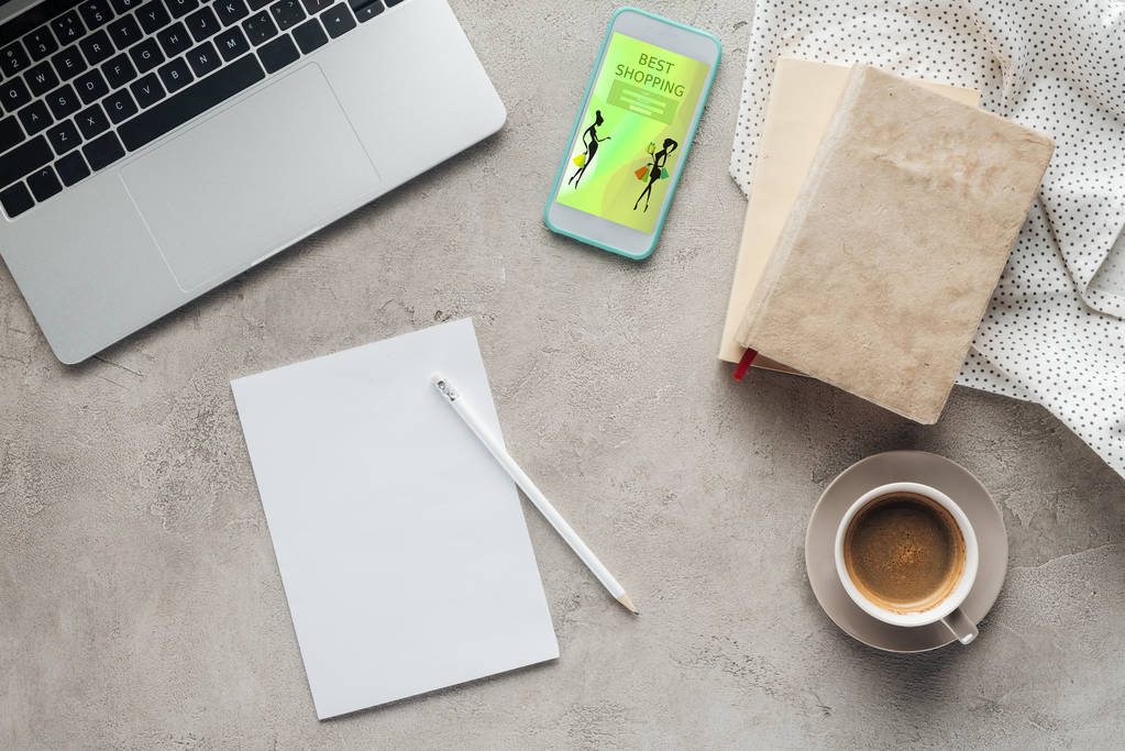 bovenaanzicht van koffie met laptop en smartphone met beste shopping app op scherm op betonnen oppervlak met blanco papier - Foto, afbeelding