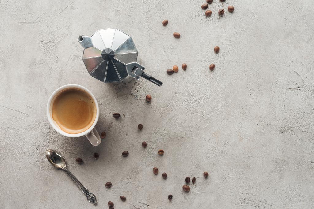Draufsicht auf Tasse Kaffee und Mokkakanne auf Betonoberfläche mit verschütteten Kaffeebohnen - Foto, Bild