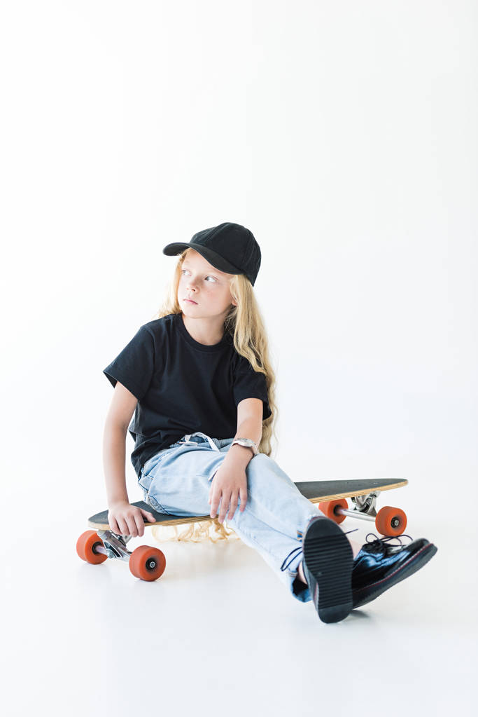 adorable child with long curly hair sitting on longboard and looking away isolated on white   - Photo, Image
